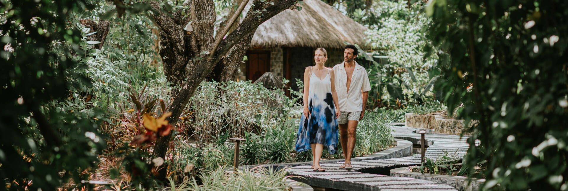 A couple walks along a winding stone path through a dense tropical garden toward a thatched-roof villa