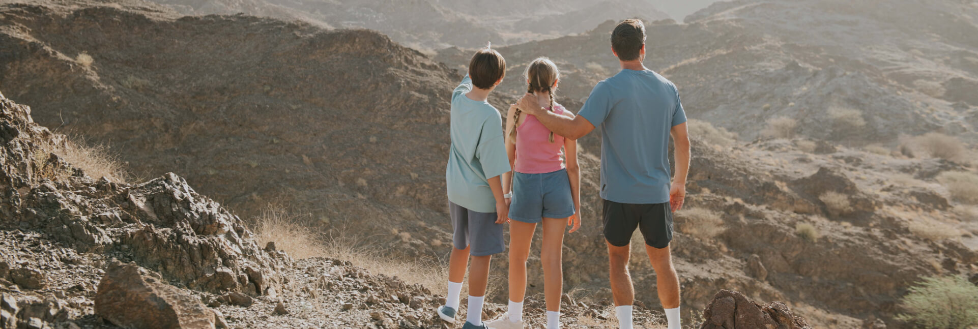 A man and two children stand on a rocky ridge, looking out over a vast, sunlit mountain range